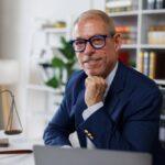 Portrait of an attractive, successful, influential, Law adult caucasian manager, businessman or lawyer in stylish formal suit, sitting at work desk in the office, looks at the camera friendly smiling